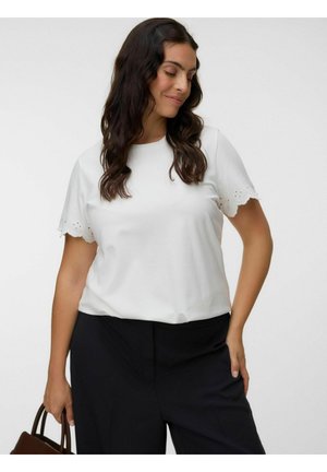 Frau mit langen dunklen Haaren trägt eine weiße Bluse mit kurzen Ärmeln und gewelltem Saum sowie eine schwarze Hose, hält eine braune Handtasche, schaut nach unten und lächelt.