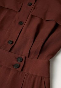 Vêtement marron avec des boutons noirs sur le devant, deux boutons sur une large ceinture à la taille et un détail de poche à rabat près de la poitrine.
