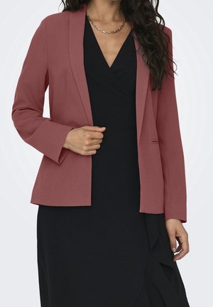 Kobieta w różowym marynarce w kolorze kurzowego różu, nosząca czarną sukienkę z dekoltem w serek, uzupełniona o złoty naszyjnik, pozująca na jednolitym tle.