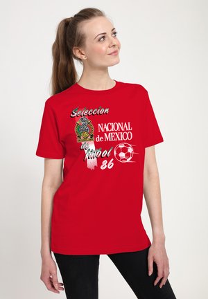 Donna che indossa una maglietta rossa con la scritta "Seleccion Nacional de Mexico Futbol 86", grafica di un pallone da calcio e un logo del calcio messicano.