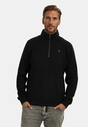 Black zip-up sweatshirt with a high collar, featuring a small red logo. Smooth fabric, long sleeves, and ribbed hem. Worn with gray jeans.