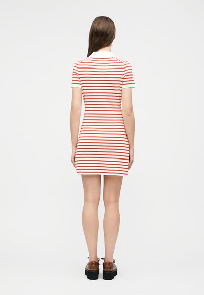 Γυναίκα που στέκεται με την πλάτη στραμμένη, φορώντας ένα κοντομάνικο φόρεμα με λευκές και κόκκινες ρίγες και καφέ πλατφόρμες, σε λευκό φόντο.