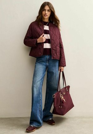 Giacca trapuntata bordeaux sopra un maglione a righe, abbinata a jeans a gamba larga blu e sneakers marroni. Borsa marrone con dettagli dorati.