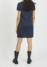 Žena s dlouhými vlnitými vlasy, oblečená v krátkých rukávech námořnického modrého šatu a černých kozačkách do půli lýtek, stojící zády k bílému pozadí.