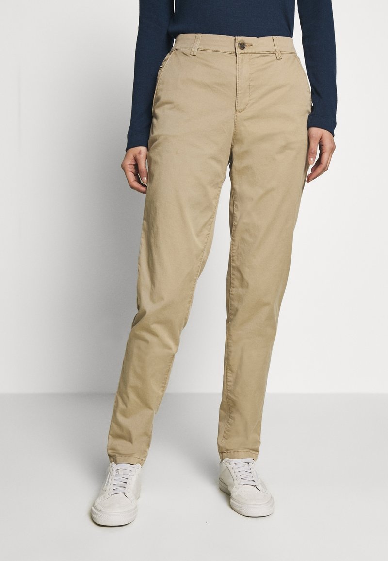 Pantaloni in cotone beige con vestibilità dritta, dotati di chiusura a bottone, tasche laterali e una texture liscia. Abbinati a sneakers bianche.
