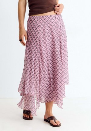 Mujer que lleva una falda midi asimétrica de cuadros rosa con sandalias marrones con tachuelas y una camiseta sin mangas de color marrón oscuro.
