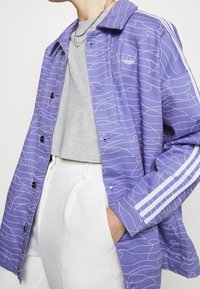 Lila jacka med vit vågmönster, knäppning med knappar och vita ränder nedanför ärmarna. Bärs över en grå topp, ihop med ljusa byxor.