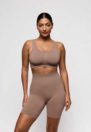 Vrouw staat zelfverzekerd, gekleed in een bijpassende naadloze bruine sportbeha en een high-waisted biker shorts tegen een effen lichte achtergrond.