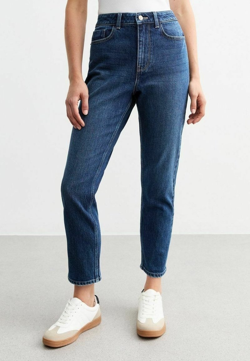 Femme portant un jean droit taille haute bleu foncé et des baskets blanches, debout sur un sol gris clair devant un mur blanc uni.
