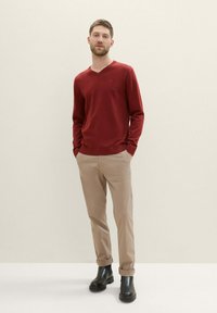Raudonas V kaklo megztinis, smėlinės slim-fit kelnės, juodi kulkšninių botai. Megztinis turi lygų paviršių, o kelnės turi pasuktą kraštą.