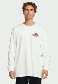 Young man with wavy hair wearing a white long-sleeve Billabong shirt and black pants, standing against a plain background.