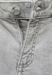 Grå denimtyg med dragkedja och tryckknappsstängning. Funktioner inkluderar sömnadsdetaljer och en blekt textur. Nära vy.