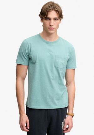 Junger Mann trägt ein hellblaues T-Shirt mit Brusttasche und schwarze Hose, steht vor einem schlichten hellen Hintergrund.