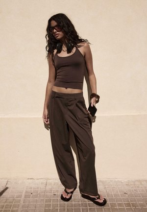 Donna con canotta marrone e pantaloni larghi posa contro un muro beige, indossa occhiali da sole, cuffie intorno al collo, tiene un telefono in mano.