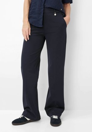 Person iført løstsiddende marineblå bukser, marineblå skjorte og sorte sneakers med hvide snørebånd, stående med den ene hånd i lommen mod en hvid baggrund.