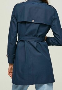 Námorně modrý trenčkot vyrobený z hladké látky, s dvojřadovým zapínáním, pásem v pase a ramenními epauletami.