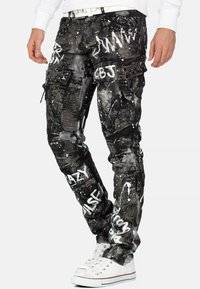 Pantalons cargo noirs et gris avec un motif de peinture éclaboussé, dotés de zips, de clous et de multiples poches. Associés à des baskets blanches.