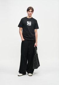 Μαύρη βαμβακερή μπλούζα t-shirt με γραφικό τετράγωνο λογότυπο, συνδυασμένη με χαλαρές μαύρες παντελόνες και λευκά αθλητικά παπούτσια. Μια μαύρη τσάντα ώμου κρατείται στο αριστερό χέρι.