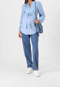 Světle modrá těhotenská blůza s detailními knoflíky a zavázáním v pase, spárovaná s džínami a bílými teniskami. Součástí je malá ramenní taška.