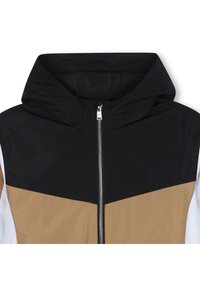 Gobtu su užtrauktuku striukė su juoda viršutine dalimi, ruda geometrine vidurine dalimi ir baltais apatine plokštėmis. Priekį puošia sidabrinė užtrauktuko linija.