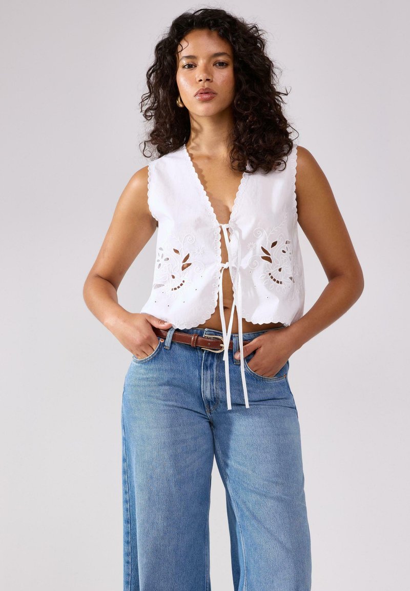Top bianco senza maniche con bordi a scallop e ricamo floreale, abbinato a jeans in denim blu a vita alta e una cintura marrone.
