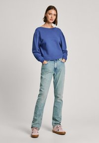 Zila krēkls ar platu kakla izgriezumu un pūkainām piedurknēm, apvienots ar gaiši ziliem džinsiem un rozā sporta apaviem. Gluda tekstūra, ikdienišķs griezums.