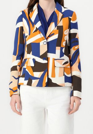 Femme portant un blazer ajusté avec un motif géométrique abstrait en bleu, orange, blanc et marron, associé à un pantalon blanc.