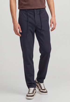 Homme portant un pantalon bleu marine foncé à taille élastique avec cordon et des baskets marron et blanches, debout devant un fond clair uni.