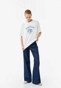 Witte t-shirt met een blauwe "Sardine" grafiek en blikontwerp, gecombineerd met donkerblauwe flared jeans en beige instapschoenen.
