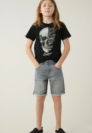 T-shirt noire avec un graphisme de crâne et du texte, associée à un short en denim léger gris clair et des chaussures noires à enfiler.
