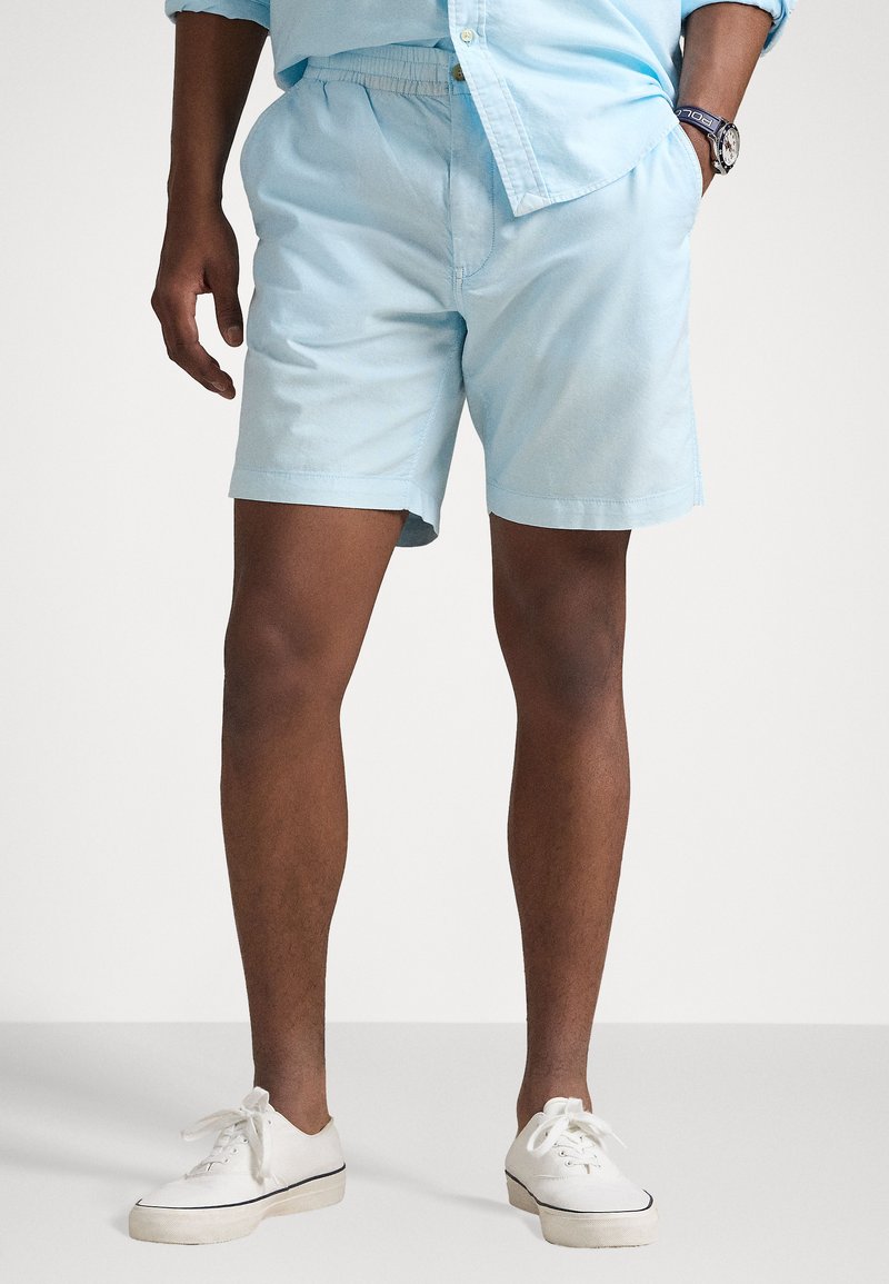 Shorts bleu clair en tissu doux et léger, dotés d'une taille élastique et de poches latérales. Portés avec des baskets blanches.