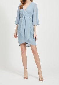 Žena v lehké modré zavinovací šatech ke kolenům se třičtvrtečními rukávy a páskem v pase, doplněná zlatými sandály na otevřenou špičku.