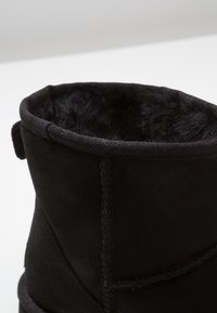 Botín de ante negro con un forro suave y peludo, detalles de costura y un puño doblado. Ausencia de herrajes o patrones visibles.