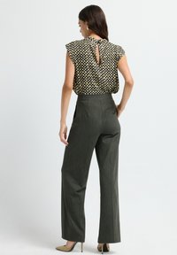 Femme portant un haut sans manches à carreaux et un pantalon large gris taille haute, se tenant avec une main sur la hanche, tournant le dos.