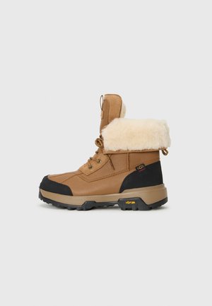 Brune vinterstøvel med en fyldig hvit pelskant og svart såle, med snøring og en løftestropp for enkel påkledning. Vibram-logo synlig.