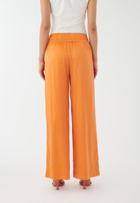 Breda byxor i vibrerande orange tyg, med elastisk midja och en slät yta. Accentuerade av smala klackar.