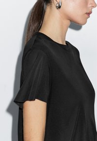 Sort T-shirt med korte ærmer og glat tekstur, rund halsudskæring og diskret bølgende ærmedetalje. Indeholder et sideprofilbillede af en model.