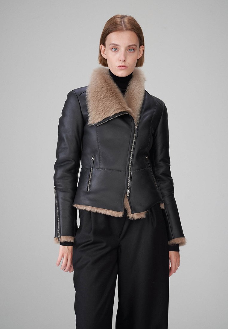 Schwarze Lederjacke mit einem felligen beigen Kragen und Saum, mit silbernen Reißverschlüssen an der Vorderseite und den Ärmeln. Glatte Textur mit taillierter Passform.