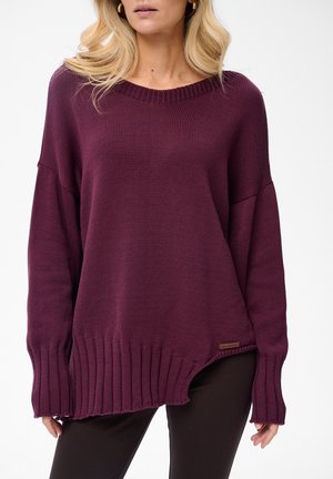 Kobieta ubrana w luźny, asymetryczny fioletowy sweter z wełny, z prążkowanymi ściągaczami i dołem, w połączeniu z czarnymi spodniami i złotymi kolczykami w kształcie kół.