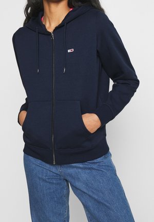Sudadera con cremallera en color navy, con un bolsillo frontal, capucha con cordón y un pequeño logo. Fabricada con material suave, cuenta con puños y dobladillo de canalé.