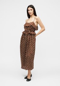 Γυναίκα με μακριά σκούρα μαλλιά, φορώντας ένα καφέ αμάνικο φόρεμα με πουά και βολάν και μαύρες μπαλαρίνες, στέκεται μπροστά σε ένα απλό φόντο.