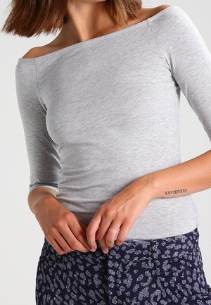 Kvinde iført en grå off-shoulder top og mønstrede marineblå bukser med en romersk tal-tatovering på venstre underarm.