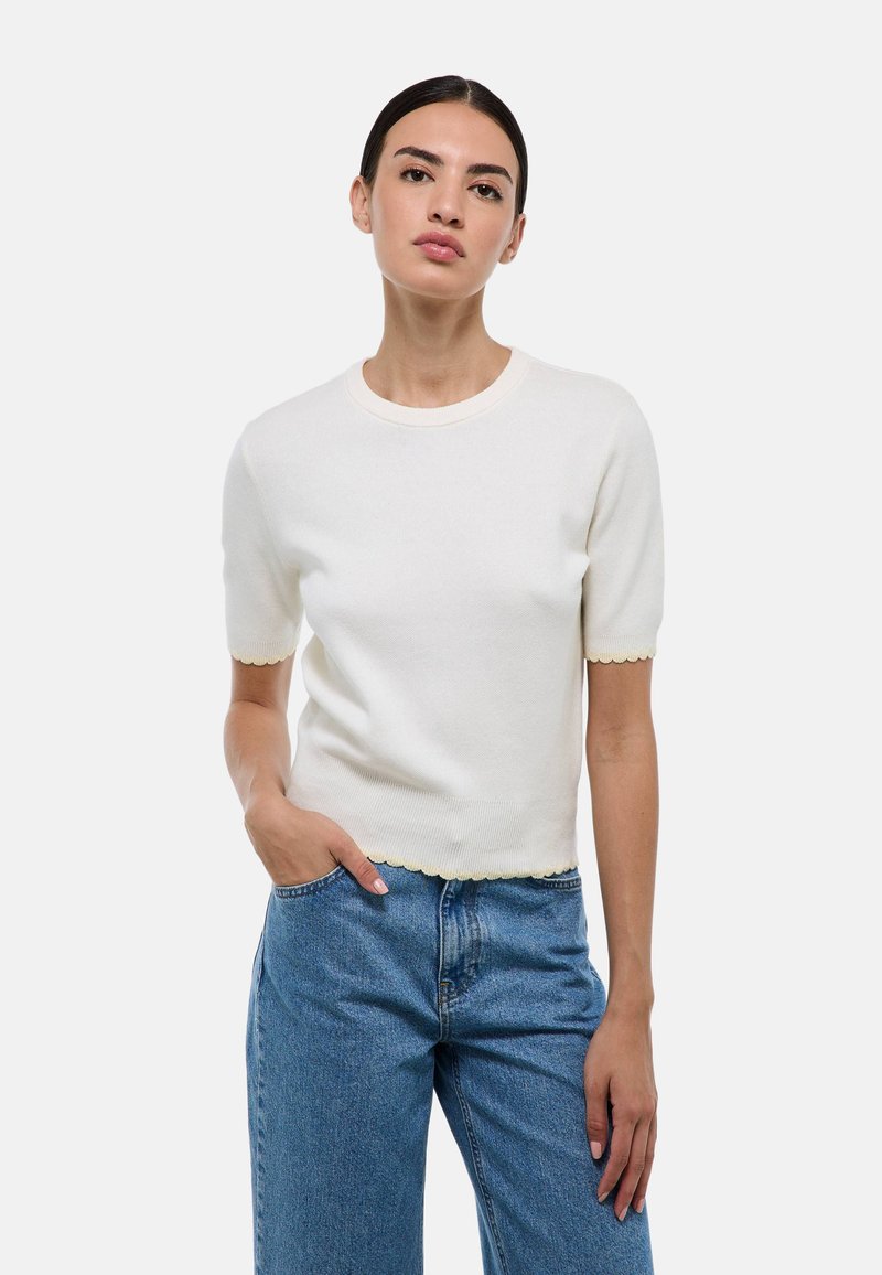 Pull crème à manches courtes avec un ourlet et un décolleté festonnés ; texture tricot douce, design ajusté, associé à un jean bleu taille haute.