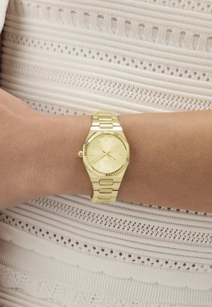 Reloj de pulsera dorado con correa de metal, usado en una muñeca izquierda sobre una tela de punto blanca texturizada.