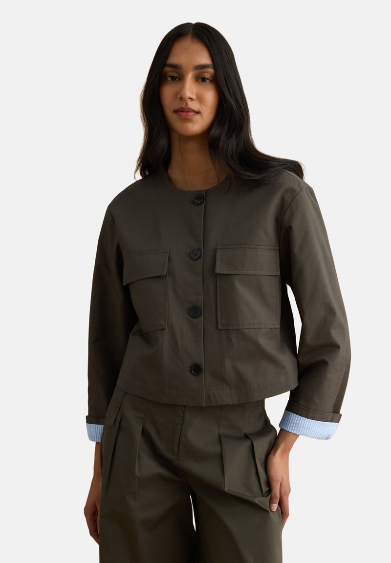 Mujer con cabello largo y oscuro que lleva una chaqueta de color verde oliva con botones y bolsillos delanteros, y pantalones plisados a juego, de pie con una mano en el bolsillo.