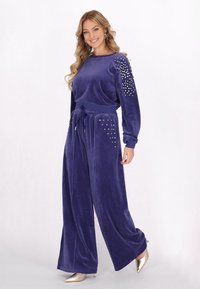 Lila velour lounge-set som består av vida byxor och en långärmad topp med strassdekorationer på axlarna och fickorna.