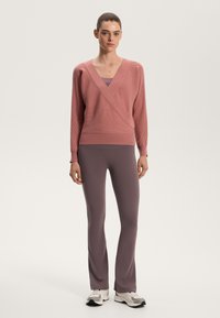 Pink knit sweater with a V-neck and crossover design, paired with form-fitting dark purple leggings. Model stands against a neutral background.