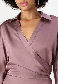 Blouse en satin rose poussiéreux, avec un décolleté en V, un col et un léger éclat. Présente des drapés subtils et des coutures nettes.
