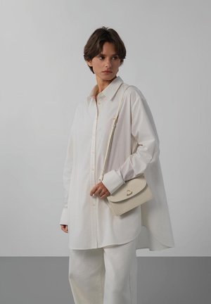 Vrouw met kort donker haar, gekleed in een oversized wit shirt en broek, met een beige crossbodytas met hartvormige gesp, staand tegen een grijze muur.