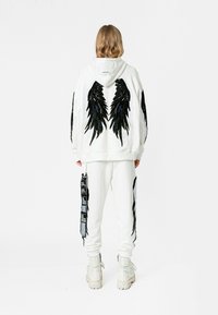 Sudadera blanca con diseños de alas negras en la parte posterior, combinada con pantalones a juego que presentan etiquetas laterales y acentos. Tejido texturizado y corte relajado.